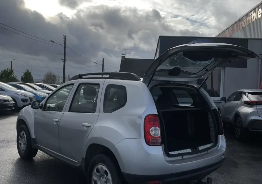 Vue 3/4 arrière droite d'un Dacia Duster gris 2011 avec hayon ouvert et attelage visible sur parking humide.