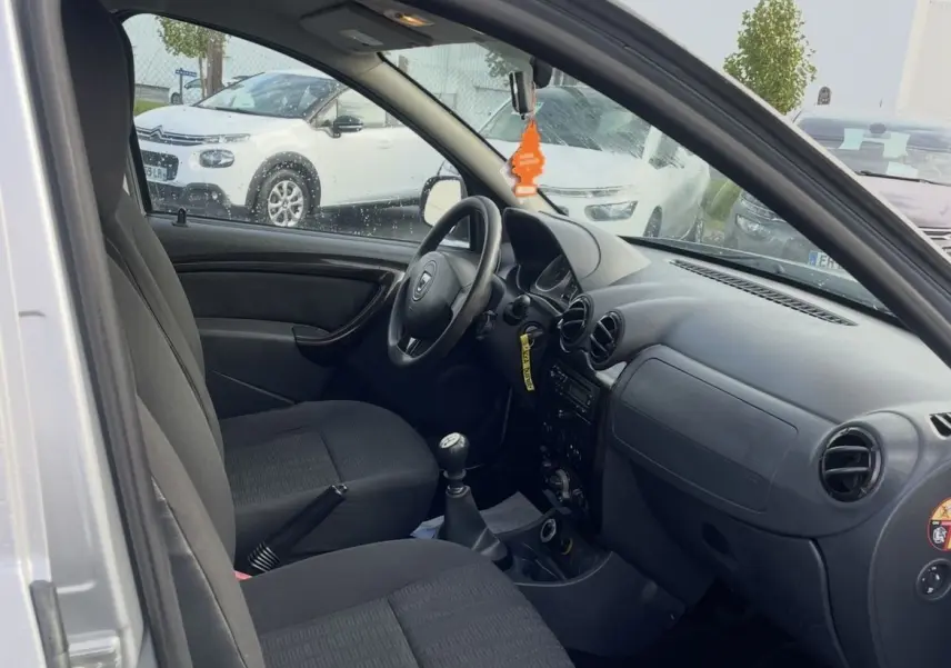 Vue intérieure côté conducteur du Dacia Duster gris 2011, montrant les sièges noirs et le tableau de bord simple avec boîte manuelle.