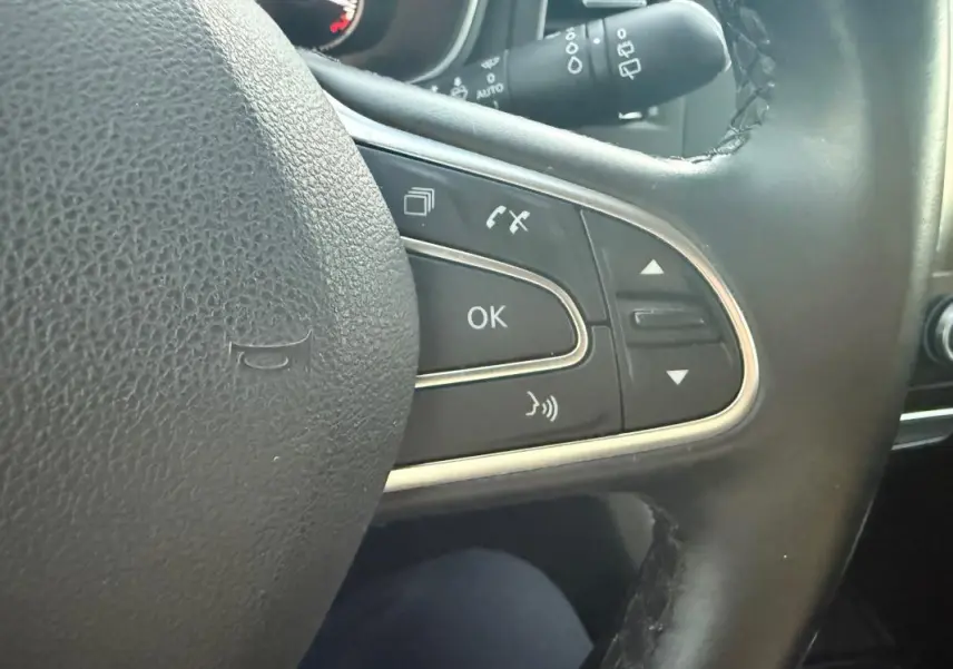 Gros plan sur les commandes du volant cuir noir de la Renault Mégane IV Berline gris clair, avec boutons multifonctions et chrome.