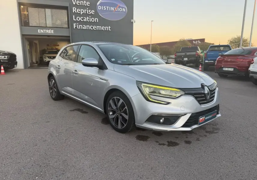 Renault Mégane IV gris clair vue 3/4 avant droit, avec phares allumés et jantes alliage 18 pouces visibles.