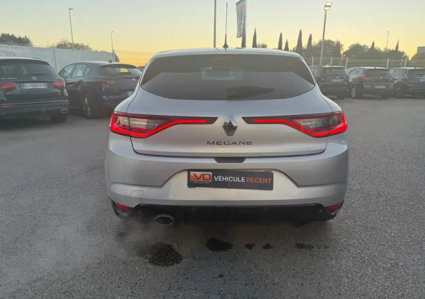 Vue arrière d'une Renault Mégane IV berline gris clair avec feux arrière allumés sur un parking extérieur.