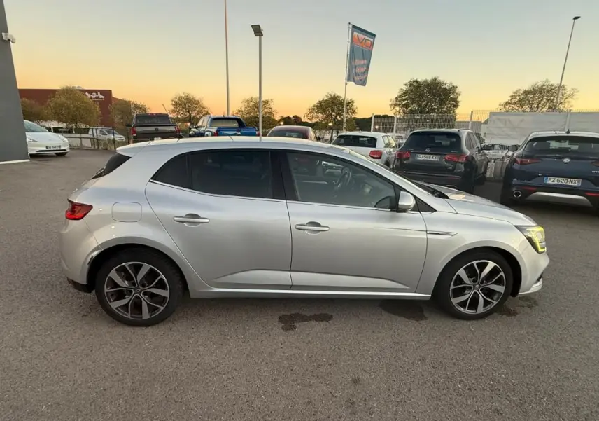 Profil côté gauche d'une Renault Mégane IV gris clair, avec jantes alliage 18 pouces, garée en extérieur au crépuscule.