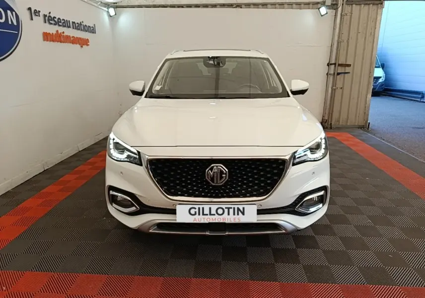 Vue frontale d’un MG Motor EHS blanc 2023 avec calandre noire et phares LED allumés dans un garage.