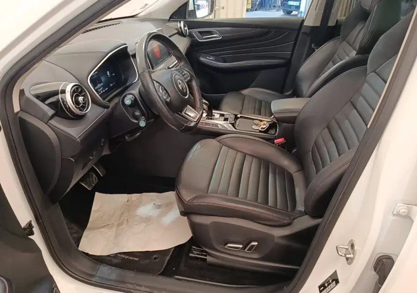 Vue intérieure côté conducteur du MG Motor EHS blanc 2023, sièges cuir noirs avec surpiqûres rouges et tableau de bord digital.