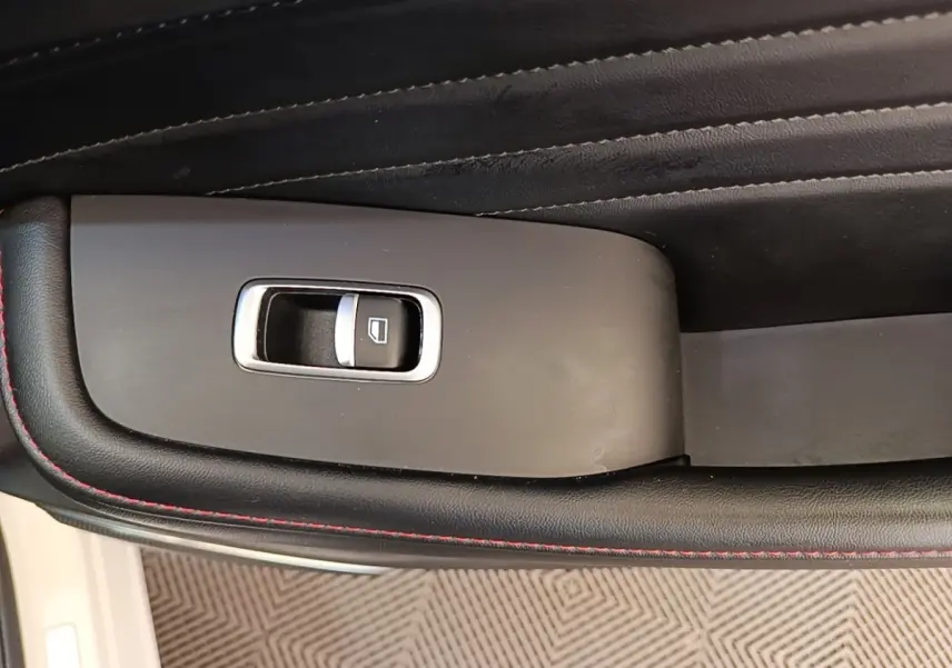 Gros plan sur le bouton de commande de la vitre électrique côté conducteur, entouré de cuir noir avec surpiqûres rouges.