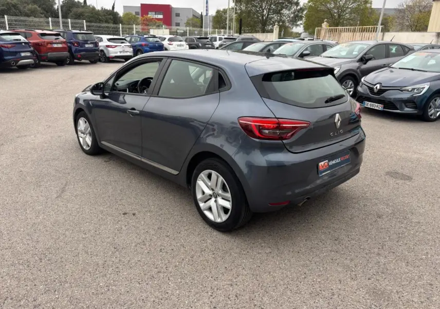 Renault Clio gris foncé vue 3/4 arrière côté gauche, avec feux arrière allumés sur parking extérieur.