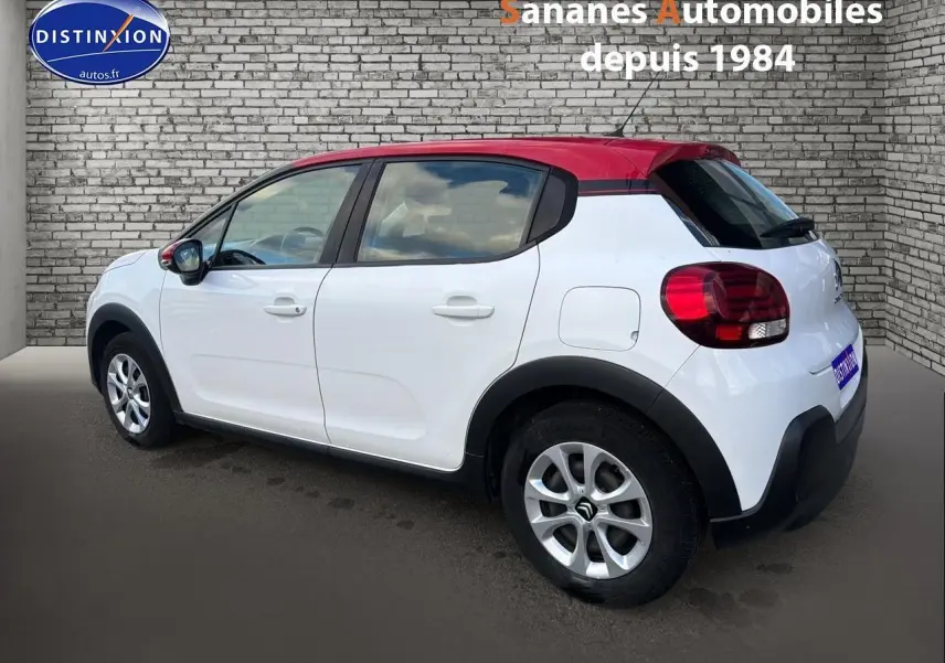 Vue 3/4 arrière droite de la Citroën C3 blanche avec toit rouge et jantes argentées, stationnée devant un mur en briques.