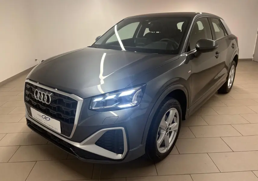 Audi Q2 35 TFSI S Line gris Daytona en 3/4 avant droit avec phares allumés dans un intérieur neutre.