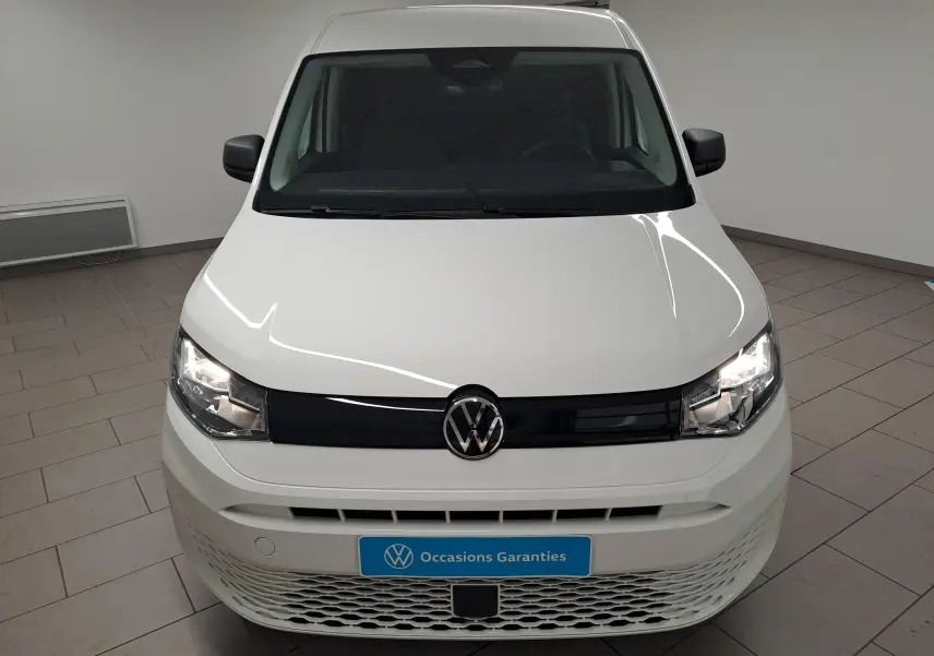 Vue avant d’un Volkswagen Caddy Cargo Maxi blanc candy 2026 avec calandre noire et phares allumés en intérieur showroom.