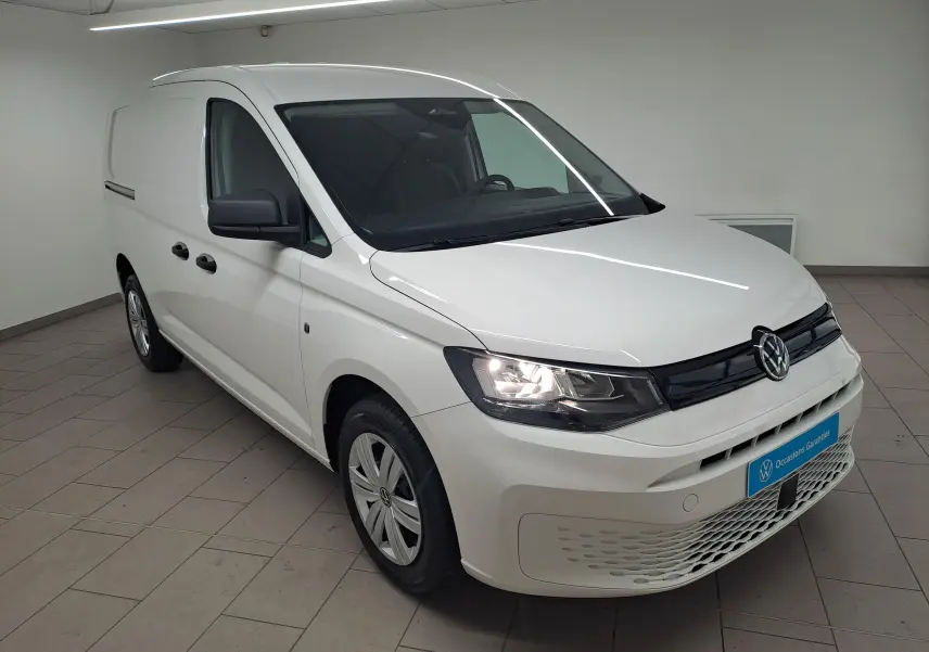 Vue 3/4 avant d'un Volkswagen Caddy Cargo Maxi blanc candy 2026 avec phares allumés en intérieur showroom.
