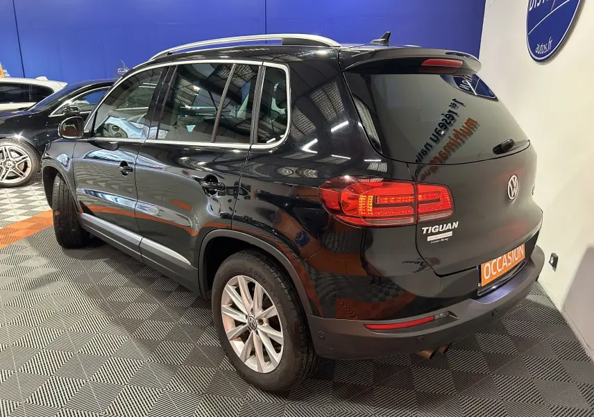 Volkswagen Tiguan noir métal vu en 3/4 arrière droit, avec feux arrière allumés et toit ouvrant visible.