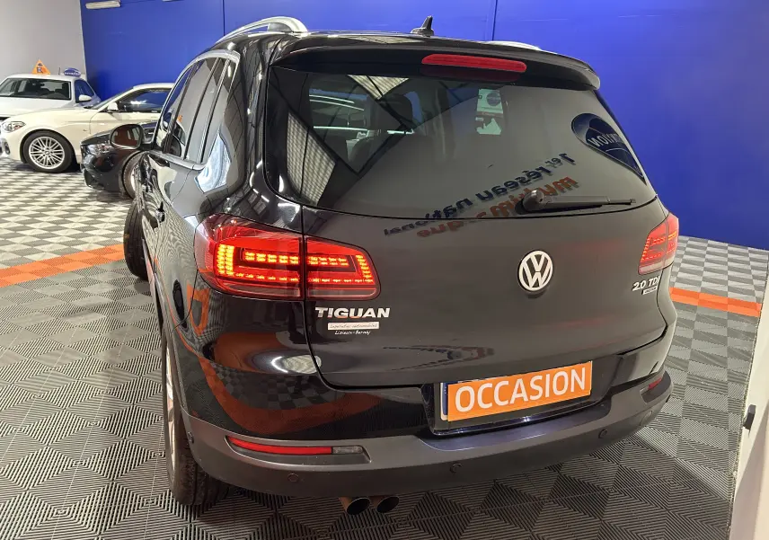 Vue 3/4 arrière d'un Volkswagen Tiguan noir métal avec feux arrière allumés et plaque "OCCASION" en intérieur showroom.