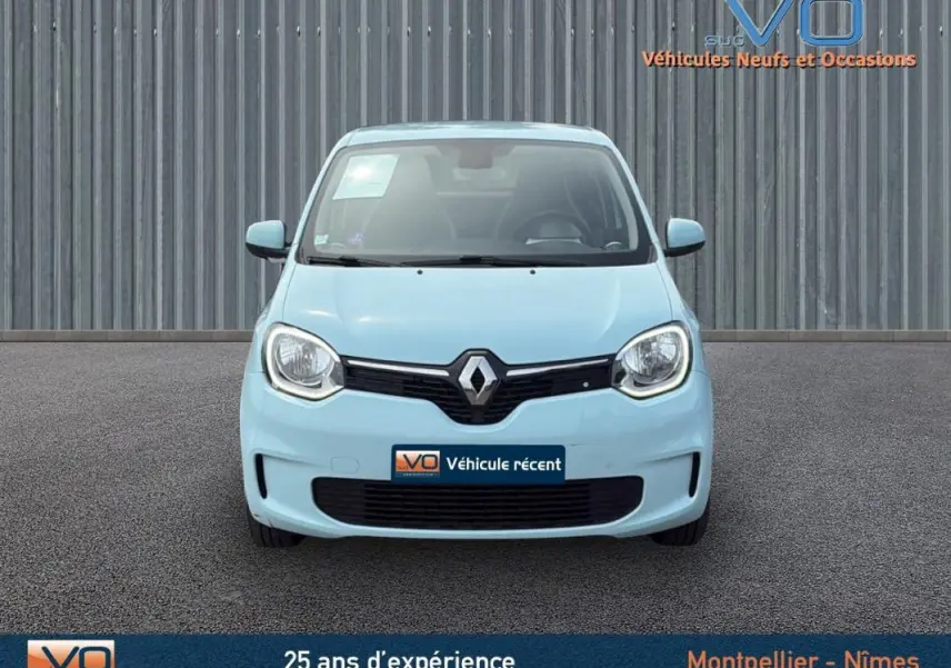 Renault Twingo III bleu clair vue de face, avec logo central et phares ronds allumés sur fond industriel.