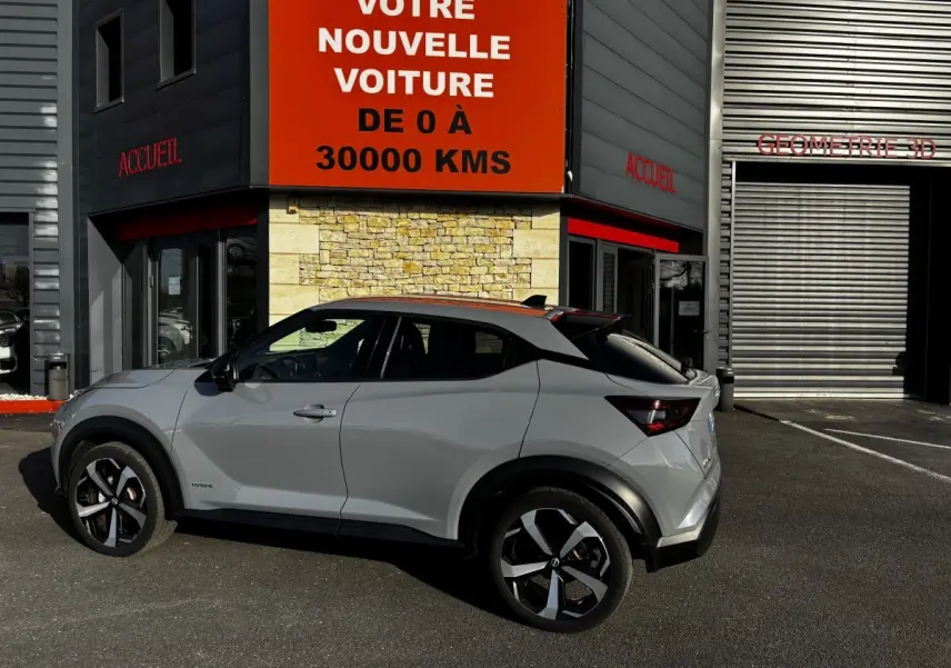 Vue de profil côté gauche d’un Nissan Juke Hybrid gris clair 2024 garé devant un bâtiment avec panneau orange.