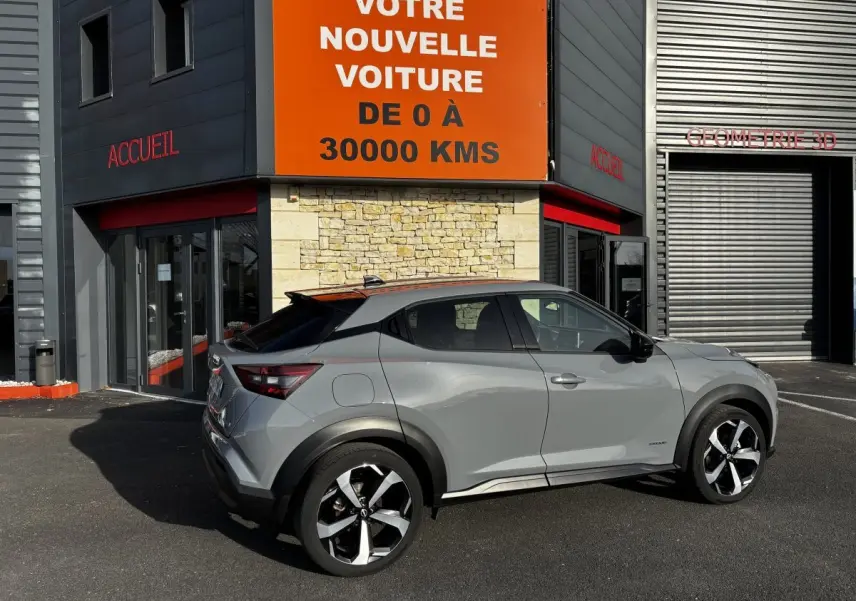 Vue de profil droit d'un Nissan Juke Hybrid gris clair avec toit noir, garé devant un bâtiment commercial.