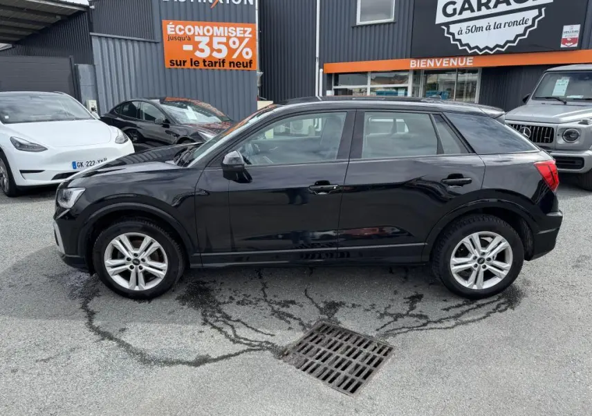 Audi Q2 noire vue de profil côté gauche, garée devant un garage avec panneaux publicitaires visibles.