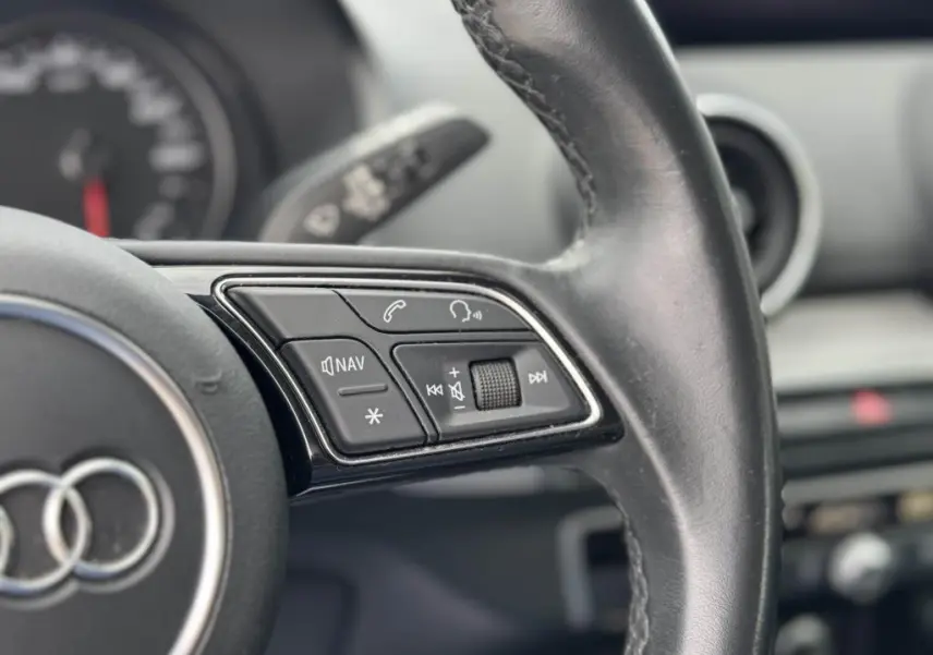 Gros plan sur les commandes gauche du volant cuir noir de l'Audi Q2 2022, avec tableau de bord flou en arrière-plan.