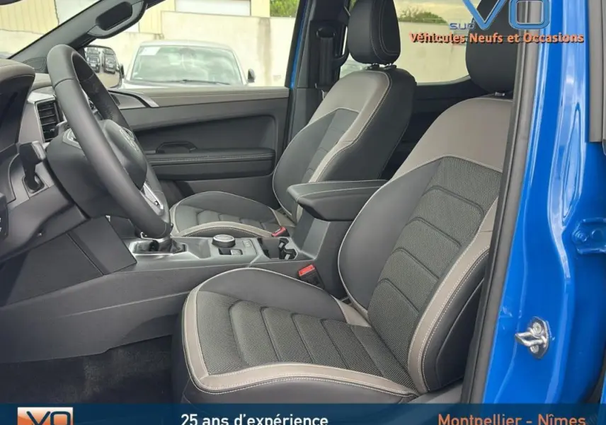 Intérieur côté gauche du Volkswagen Amarok Aventura bleu 2025, montrant sièges cuir gris avec accoudoir central et volant multifonctions.