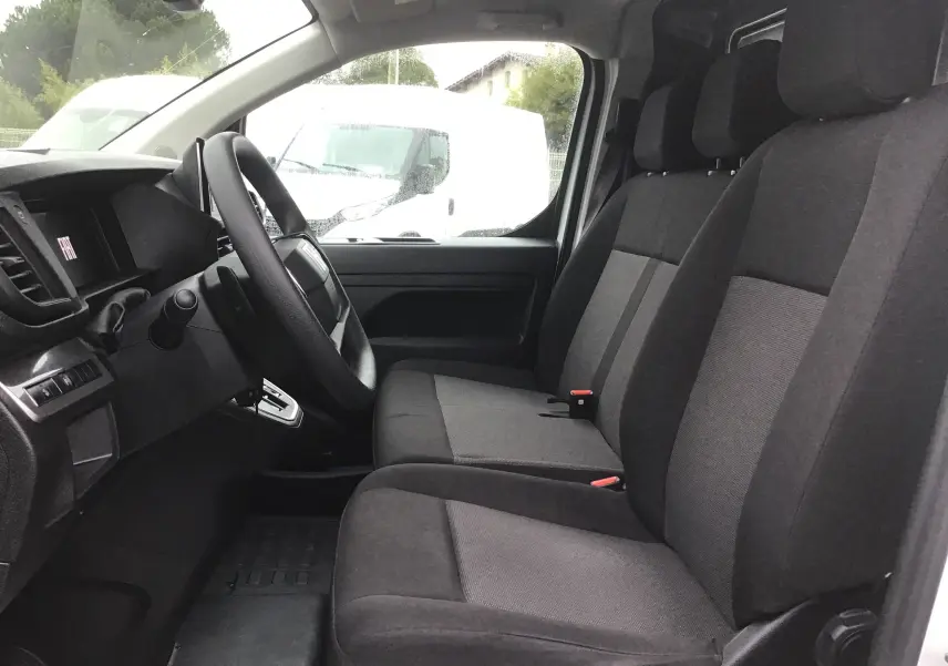 Vue intérieure côté conducteur du Fiat Scudo Fourgon blanc 2024, montrant les sièges avant en tissu gris et le tableau de bord.