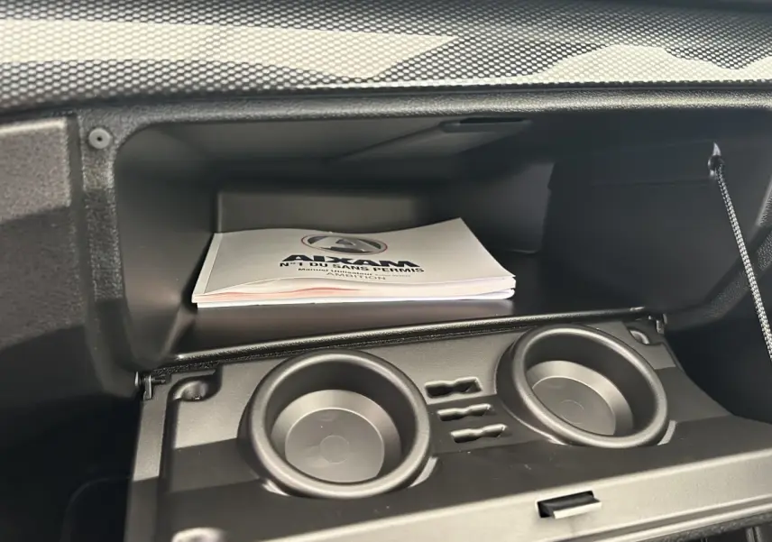 Gros plan sur la boîte à gants ouverte de l'AIXAM AMBITION blanche, avec porte-gobelets intégrés et manuel visible.