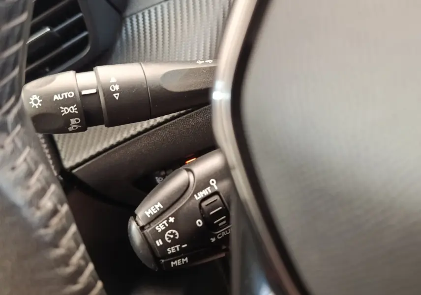 Gros plan sur les commandes de régulateur de vitesse et éclairage derrière le volant cuir d'une Peugeot 208 blanche.