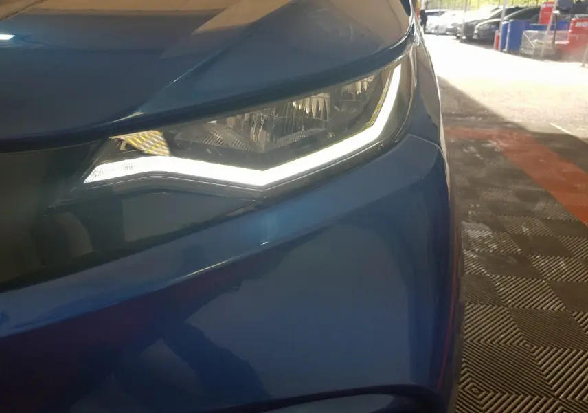 Gros plan sur le phare avant gauche allumé de l’AIXAM AMBITION 0.5 CITY SPORT bleu 2026, avec carrosserie brillante.