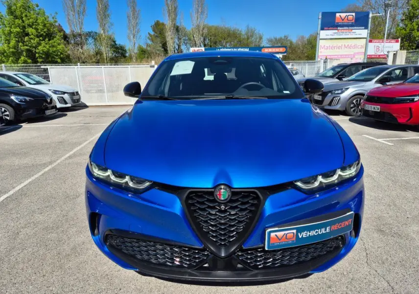 Vue avant d'une Alfa Romeo Tonale bleu vif avec calandre distinctive et feux LED allumés, en extérieur sur parking.