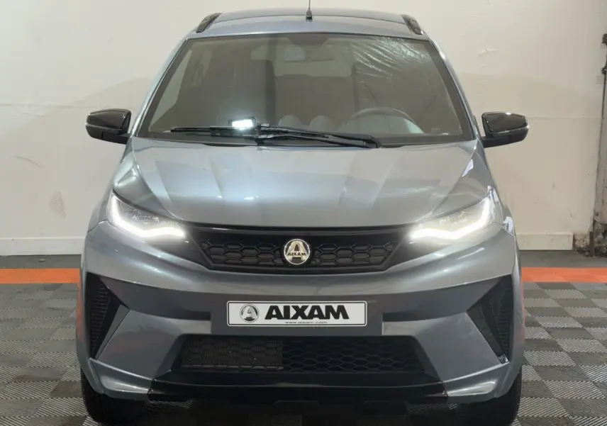 Vue frontale d'un Aixam Ambition 0.5 Crossover Premium gris foncé avec phares allumés et rétroviseur caméra côté gauche.