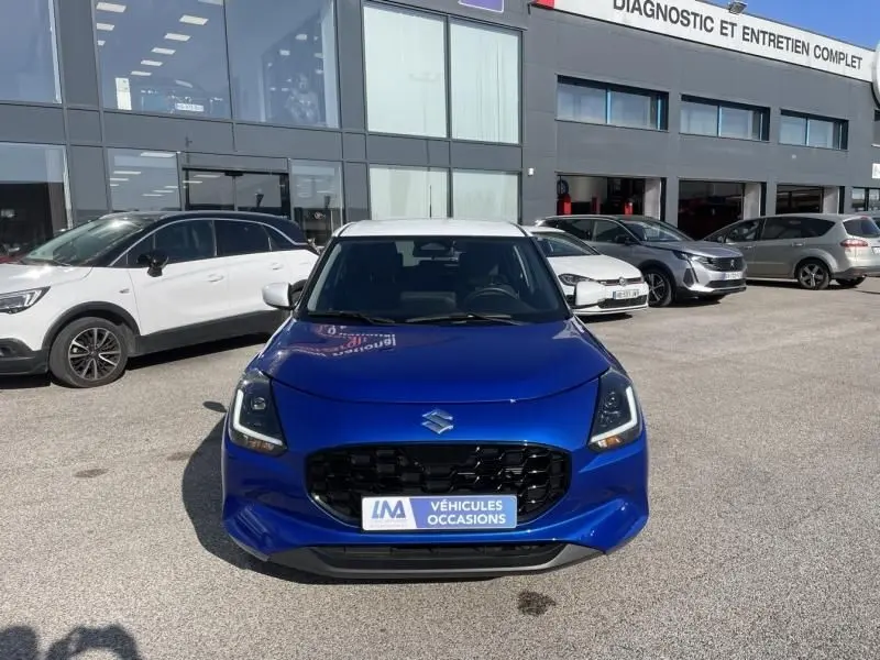 Suzuki Swift 1.2 Dualjet Hybrid bleu métal vue de face, calandre noire et toit blanc visible, devant un garage.