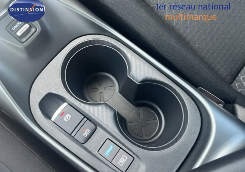 Gros plan sur la console centrale du Nissan Juke Hybrid gris, montrant les porte-gobelets et les boutons de contrôle électrique.