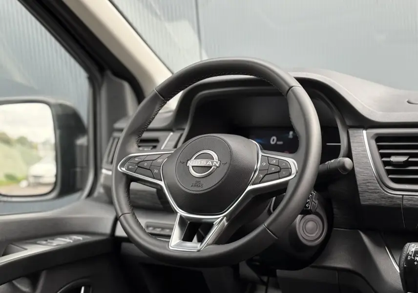 Volant Nissan en cuir avec commandes intégrées, tableau de bord noir et rétroviseur côté gauche visible.
