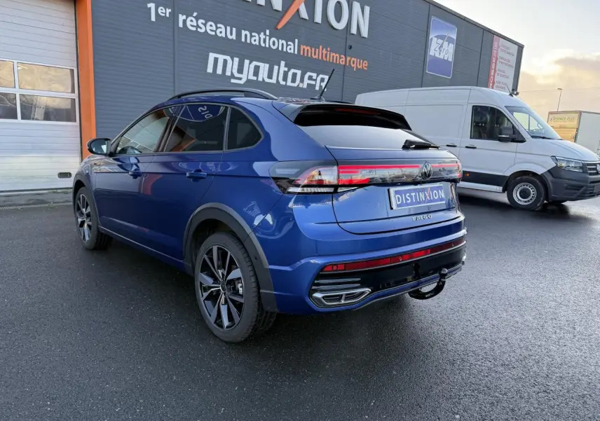 Vue 3/4 arrière droite du Volkswagen Taigo bleu récif métallisé avec jantes noires brillantes et attelage amovible.