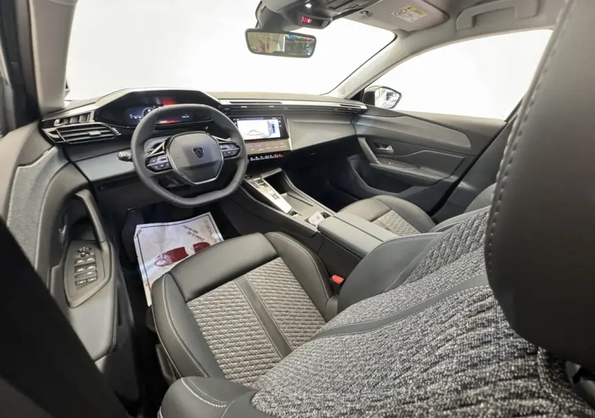 Intérieur de la Peugeot 308 2026 vu côté conducteur, sièges cuir et tissu gris, tableau de bord avec écran tactile.