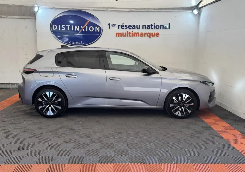 Profil droit d'une Peugeot 308 gris clair 2026 dans un showroom avec jantes noires et logo Distinxion en arrière-plan.
