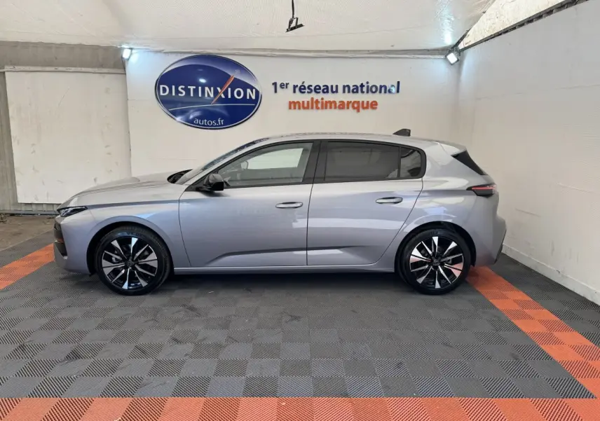 Vue de profil côté gauche d'une Peugeot 308 gris clair 2026 dans un showroom avec jantes noires et logo Distinxion en arrière-plan.