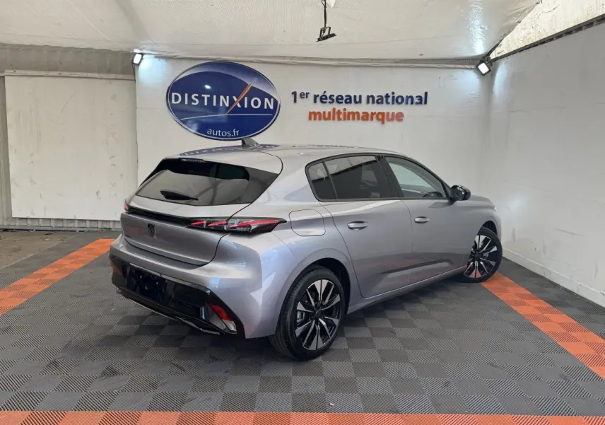 Vue 3/4 arrière droite d'une Peugeot 308 gris clair 2026 dans un showroom avec jantes noires et feux LED distinctifs.