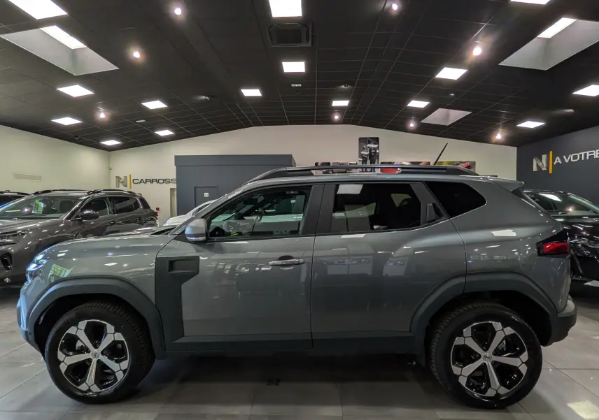 Vue de profil côté gauche du Dacia Duster Hybrid-G 150 CH 4x4 gris schiste avec jantes bi-ton et barres de toit en showroom.