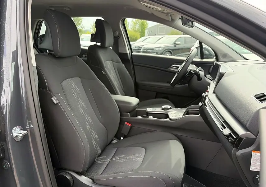 Vue intérieure côté conducteur du Kia Sportage 1.6 hybride gris eclipse, sièges tissu noirs avec surpiqûres blanches et console centrale moderne.