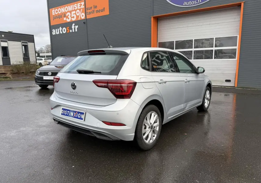 Volkswagen Polo 2025 couleur reflet d'argent métallisé vue 3/4 arrière droit sur parking humide devant un garage autos.fr