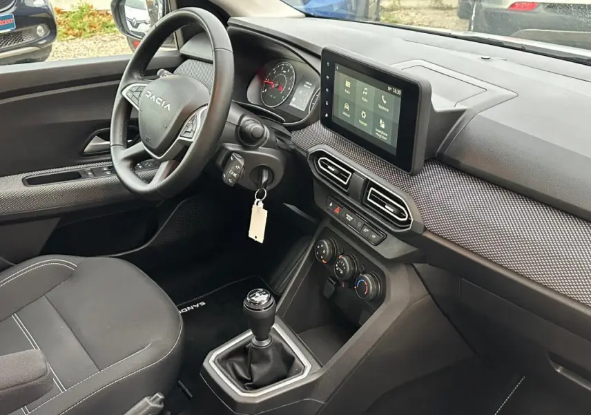 Vue intérieure côté conducteur de la Dacia Sandero blanche 2024, volant, écran tactile et levier de vitesse manuel visibles.