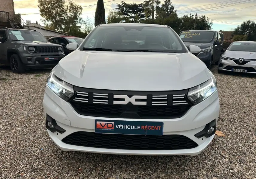 Vue frontale d'une Dacia Sandero blanche 2024 avec phares allumés et calandre noire distinctive.