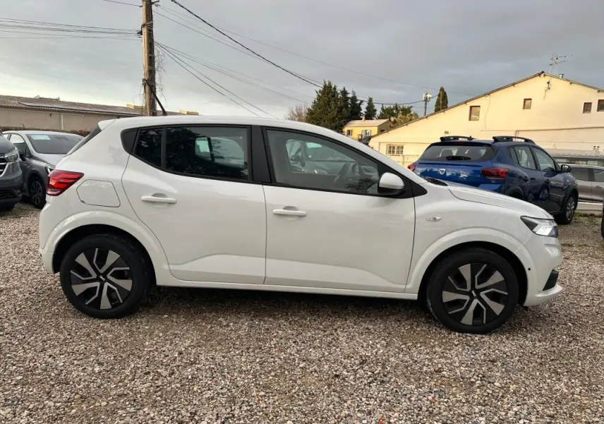 Vue de profil côté gauche d'une Dacia Sandero blanche 2024 garée sur un sol gravillonné, avec jantes noires et argentées.