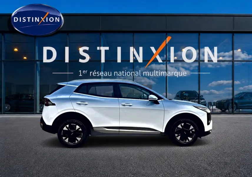 Profil droit d'un KIA SPORTAGE 1.6 hybride 239 blanc, avec jantes alu 17 pouces, devant une vitrine Distinxion.