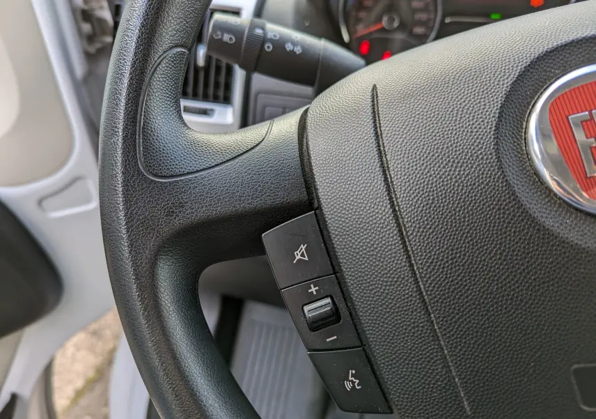 Gros plan sur le volant noir du Fiat Ducato Fourgon blanc, avec commandes audio intégrées et logo FIAT visible.