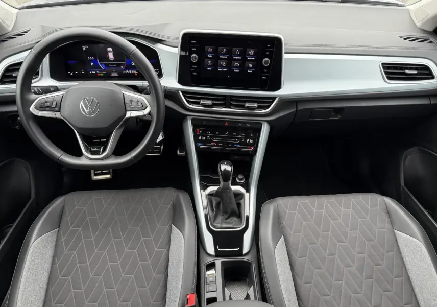 Vue intérieure avant du Volkswagen T-Roc 2025, tableau de bord moderne avec écran tactile et sièges tissu gris à motifs géométriques.