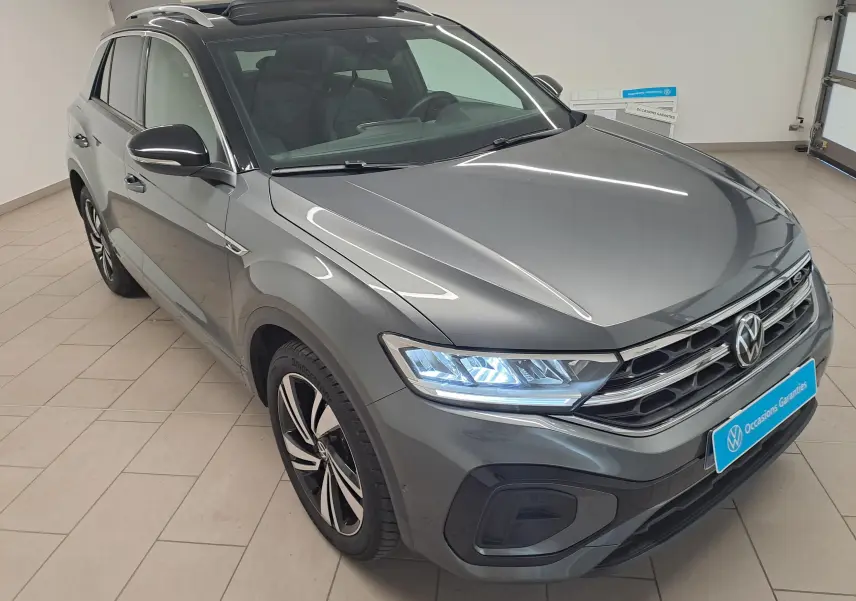 Vue 3/4 avant droite d’un Volkswagen T-Roc 2025 gris Indium avec toit noir et phares LED allumés dans un showroom.