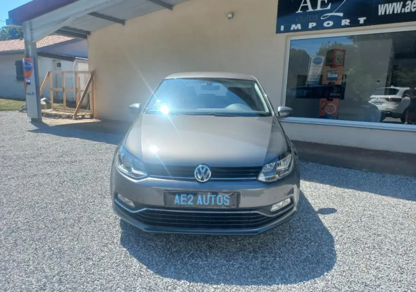 Vue de face d'une Volkswagen Polo gris métallisé 2016, avec plaque AE2 AUTOS, stationnée devant un bâtiment.