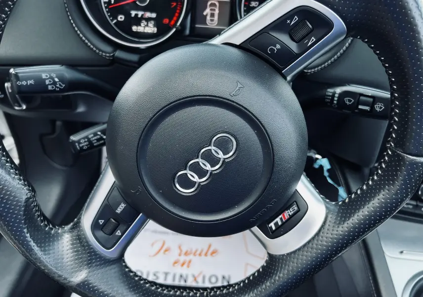 Volant en cuir perforé avec logo Audi au centre, tableau de bord et compte-tours TT RS visibles en arrière-plan.