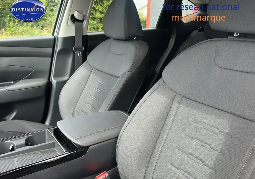 Gros plan sur les sièges avant gris du Hyundai Tucson 2025, mettant en valeur l'accoudoir central et la ceinture de sécurité côté gauche.