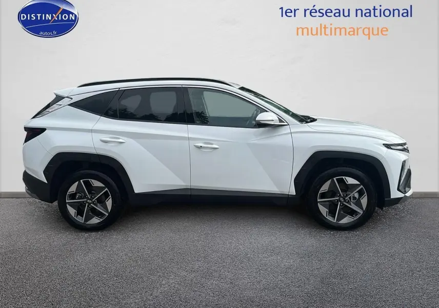 Profil côté gauche du Hyundai Tucson 2025 blanc sérénité avec jantes alliage 18 pouces et barres de toit noires.